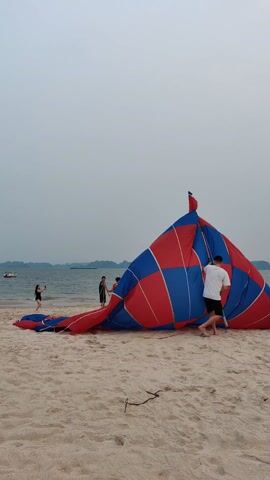 Here's a description of the video:

On a sandy beach under an overcast sky, a large, deflated parachute or balloon with a red and blue checkerboard pattern lies on the sand. A man wearing a white shirt and dark shorts stands beside the parachute, seemingly adjusting or preparing it. He then picks up a segmented pole. In the background, a woman stands at the water's edge, appearing to take photos, and a small boat is visible on the water. The sea is calm, and distant islands are visible on the horizon. The beach appears empty except for the two people.