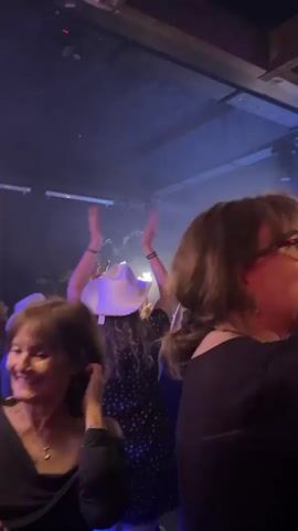 The video shows a group of people dancing at what appears to be a crowded party or club. The room has a low ceiling with exposed beams, and the lighting is dark with blue and red stage lights. Several people are holding up sparklers and cell phones, and the atmosphere seems lively and celebratory. The focus shifts between different individuals in the crowd, highlighting their reactions and movements. Many people are wearing cowboy hats.
