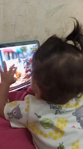 A young child lies on their stomach, watching a tablet computer. The tablet displays images of cats and babies. The child wears a white t-shirt with a yellow and green pattern. The background is a textured wall.