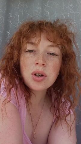 A close-up shot captures a woman with curly red hair and freckles, wearing a pink top and a necklace with a cross pendant. She is speaking directly to the camera. The background is a muted gray curtain with a textured pattern.