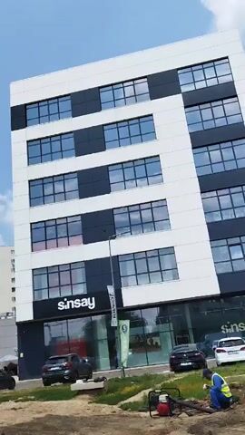 The video shows a street view of a modern commercial building with a Sinsay store on the ground floor. The building is multi-story with a white and dark gray facade and large windows. In front of the building, there is a bus stop sign and a traffic sign indicating a speed limit of 30. Several cars are parked in a lot in front of the building, and some people are walking on the sidewalk. The sky is blue with some white clouds, and the overall environment appears to be an urban or suburban setting. The camera pans to the right, showing more of the commercial building and apartment complexes in the background. There are also other stores visible on the ground floor, including Pepco and Mega Image. The video captures a sunny day with good visibility.