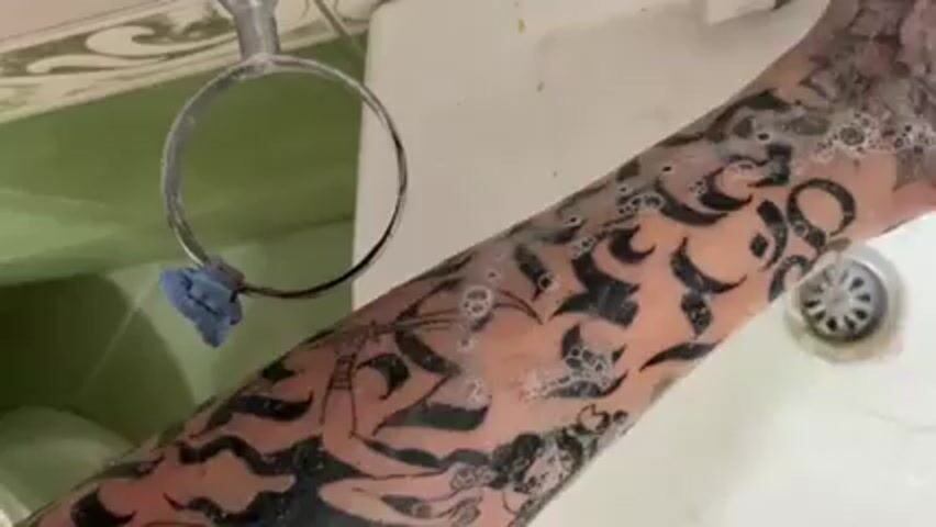A person’s forearm is being washed under a faucet. The forearm has extensive black ink tattoos with some parts obscured by soap suds. The tattoos appear to depict stylized flames and a depiction of a deity. The forearm is being scrubbed with the fingers and palm of a hand. The sink is white and has a silver drain. The faucet is chrome. The wall above the sink is green and white.