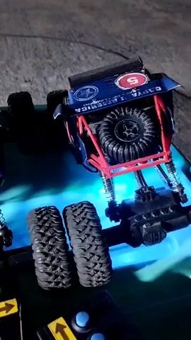 A toy rock crawler is positioned on a flat, dark surface, likely outdoors, given the background. The toy vehicle is blue and red with a roll cage. It has large, treaded tires, and a spare tire is mounted on the rear. There are two additional large tires to the left of the vehicle. A green surface, possibly a mat or a table, is beneath the toy. The scene is lit by artificial light, creating blue and green reflections on the surface. There are two remote control units in the foreground with yellow directional arrows. The video is shaky and the camera is angled downwards, focusing on the toy vehicle.