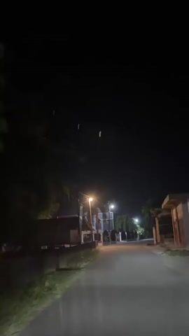 The video shows a dark, empty street at night. Streetlights illuminate the road and the sides of buildings. Trees are visible on either side of the road, with some of their leaves illuminated by the streetlights. The camera is moving, suggesting it is from a vehicle. There are no people or animals visible in the video. The sky is dark with a few faint green lights that could be distant lights or lens flares.