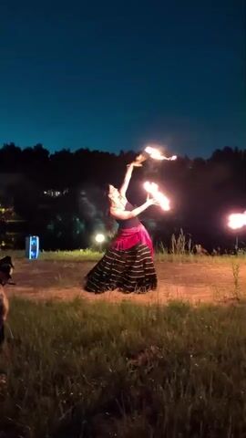 A woman in a black dress with a pink sash is performing a fire dance outdoors at night. She is holding two flaming objects, one in each hand, and appears to be spinning them. The background shows a dark sky with trees and water in the distance. There is also some grass in the foreground and a light source to the left.