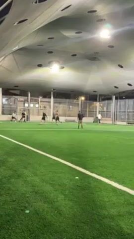On an artificial turf soccer field lit by overhead lamps, several people are engaged in a soccer game. One player, shirtless, dribbles the ball towards the goal defended by a man in black. Another player in black moves to intercept. Additional players are positioned around the goal, observing the action. A white line marks the field, and a fence runs along the perimeter.