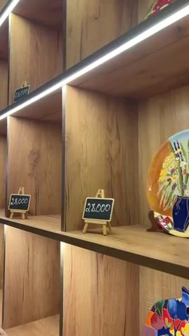 The video shows colorful painted plates displayed in an interior space. The first shot shows a wooden shelf unit with multiple compartments, each containing a painted plate and a small chalkboard with the price of "28,000" written on it. The camera pans out to show the entire wall-mounted shelf unit, which is lit by strip lighting above each shelf. A doorway is visible on the left. The camera then pans towards a wall decorated with more of these painted plates. A staircase with lights beneath the steps is also visible.