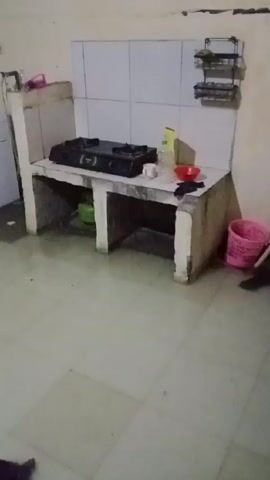 The camera pans across a cluttered kitchen. A tile-topped cooking surface holds a two-burner gas stove, some cleaning supplies, and a red bowl. A green gas cylinder is stored underneath. A white washing machine stands against the far wall. The tiled floor is dirty with scattered objects including a pink bucket, a red item, a small black object, and a discarded clear plastic bottle.