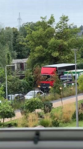 A red truck is parked on a road that curves to the right. The truck's trailer is white with green accents and text. To the left of the truck, a white car is parked. Behind the truck and to the right, other vehicles are visible, including a green truck and a blue car. Lush green trees and foliage surround the scene, and a tall electrical pole stands in the background. The sky is overcast.