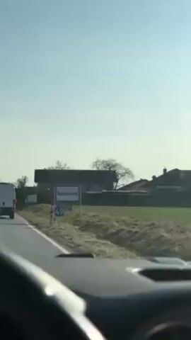 The video shows a car entering Ravensburg, Germany. A road sign with the town's name is visible on the right side of the road. Behind the sign are houses, bare trees, and a field. The sky is clear and blue. The video is shot from the driver's perspective. A white van is ahead of the car.