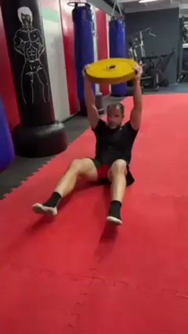 A medium-shot video shows a man performing an exercise in a gym. The man, of medium build with short dark hair, is wearing a black t-shirt, black shorts with red trim, black socks, and grey sneakers. He is sitting on a red mat that is composed of interlocking puzzle pieces, with his legs extended outwards in a V-shape. He holds a yellow weight plate overhead with both hands. He is performing seated Russian twists, rotating his torso from side to side while keeping the weight elevated. Behind him are several punching bags and exercise equipment. The punching bags are red, black, and blue. One of the punching bags has a drawing of a muscular man on it. The gym has a white ceiling with visible light fixtures. The video is shot from a stationary position. The lighting is artificial. There is some ambient noise, including shouting and cheering.