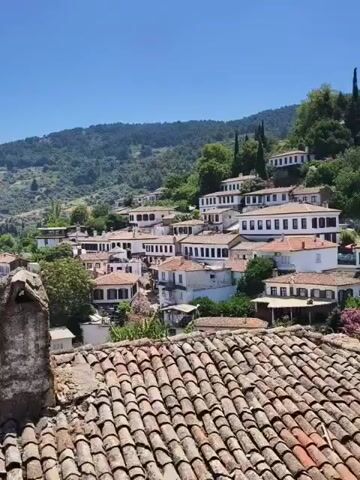 The video shows a panoramic view of a village nestled on a hillside. The houses are predominantly white with red tiled roofs, characteristic of Mediterranean or Aegean architecture. They are built close together, cascading down the slope. Lush green trees and vegetation surround the village, and a dense forest covers the higher slopes of the mountain in the background. The sky is a clear, bright blue, indicating a sunny day. In the foreground, the textured surface of more red tiled roofs is visible, suggesting the vantage point is from a higher elevation or another building. The overall impression is one of a charming, historic, and serene settlement bathed in natural light.