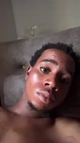 A man lies on a bed or couch, looking up at the camera. He has dark skin, short dark hair, and a thin beard. His skin appears shiny. The headboard behind him is gray and tufted. The background is a plain, off-white wall.