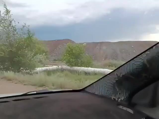 The video captures a view from inside a vehicle, looking out through the windshield. The landscape appears rural or semi-arid with trees and some man-made structures. A large, light-colored pipe or conduit runs along the side of the road, winding its way through the terrain. The sky is overcast with clouds. The vehicle appears to be in motion, causing the trees and scenery to blur slightly. The dashboard and part of the windshield are visible in the foreground. There are no people clearly visible in the frame.