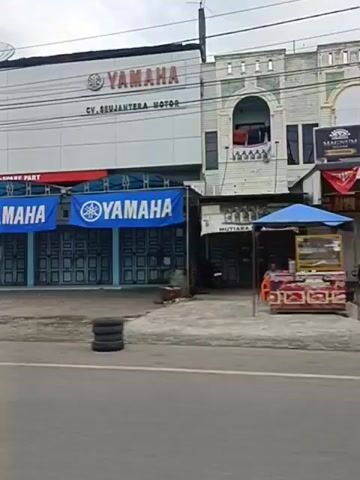 The video shows a street scene with several buildings and vehicles. On the left, there is a Yamaha motorcycle dealership with a white facade and blue banners. A yellow truck and a motorcycle are visible in front of the dealership. The street appears to be a commercial area with other businesses, including a shop with a green awning. Several vehicles are driving on the road, including a black SUV. The overall environment suggests a moderately busy urban or suburban setting.