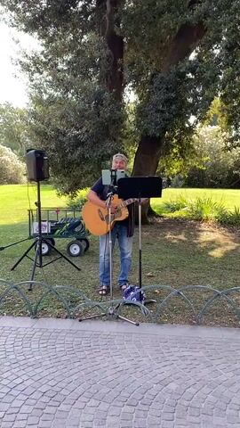 A man stands in a park playing an acoustic guitar and singing into a microphone. He is wearing a blue t-shirt, jeans, and a bandana. To his left is a speaker on a tripod, and behind him is a cart with items on it. To his right is a music stand. The man is standing on grass, and in front of him is a paved area with a decorative metal fence. Trees and bushes are visible in the background. The lighting suggests it is daytime.