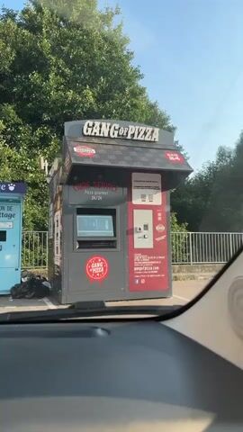 The video shows a "Gang of Pizza" vending machine. It is a dark gray and red automated kiosk with a large sign that reads "GANG OF PIZZA." The machine has a touchscreen display on the left side, a payment terminal and a slot for cash or cards on the right side, and a smaller screen above the payment terminal. There are QR codes on the right side of the machine, along with instructions in French. The text on the machine indicates that it offers "Pizza gourmet 24/24" (gourmet pizza 24/7) and "Libre Service" (self-service). The machine appears to be located outdoors, possibly in a parking lot, with trees and a blue vending machine visible in the background. The video is taken from inside a vehicle, with the dashboard visible in the foreground. The lighting suggests it is daytime.