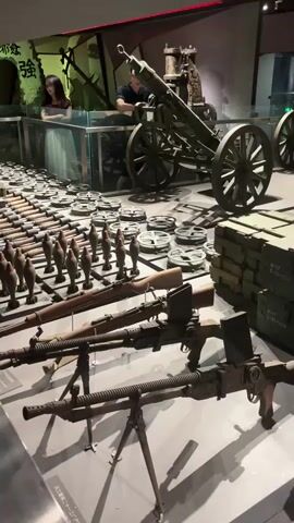 The video pans across a museum exhibit of various weapons and ammunition. In the foreground, several rifles and machine guns are displayed on stands. Behind them, rows of mortar shells are arranged, followed by stacks of ammunition crates. Further back, a large artillery piece with wheels is visible, along with more ammunition. In the background, two individuals, a woman and a man, stand behind a glass barrier, observing the exhibit. The environment appears to be a dimly lit museum hall.