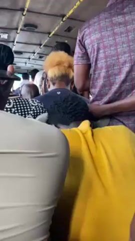 The video shows the interior of a crowded bus. Many people are standing. Passengers wear a variety of clothing. One person is wearing a yellow shirt, another a burgundy and gray plaid shirt, and another a black and white polka dot patterned top. One person has blonde hair. The bus windows are visible.