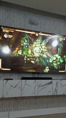 Here's a detailed description of the video:

The video shows someone playing Mortal Kombat on a large flat screen television mounted on a wall. The game is in progress, and two characters are engaged in combat in a dark, swampy environment with lush green vegetation and some kind of web or netting. The health bars of both characters are visible at the top of the screen along with other game information. The TV is mounted above a white, marble-patterned console. The wall behind the TV has a vertical ribbed design and is painted in a light grey color. There is a circular light fixture mounted on the ceiling.