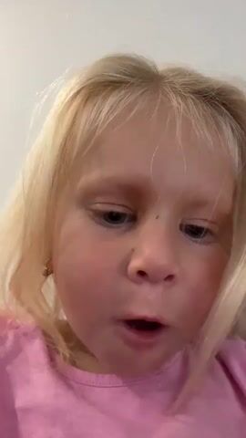 The video is a close-up, head-on shot of a young Caucasian girl with blonde hair and blue eyes. She is wearing a pink t-shirt with a design on it. Her facial expressions change throughout the video, from a neutral expression to a smile, then to a pouting or crying expression, and then back to a neutral expression. Her hands are visible at times, moving near her face and head. The background is blurred and appears to be an indoor setting, possibly a living room or bedroom. The lighting is consistent and seems to be coming from the front. There is a small dark speck on her forehead.