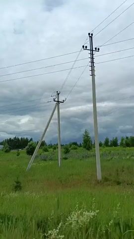 A video shows a grassy field with scattered trees and some houses in the distance under a cloudy sky. Power lines run across the top of the frame. A utility pole is visible on the right side of the frame, with another utility pole in the distance on the left. A road curves through the scene on the left. The video appears to be shot from a moving vehicle, as indicated by the side mirror visible at the bottom left of the frame.