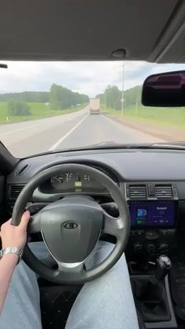 The video presents a first-person perspective from inside a car driving on a paved road under an overcast sky. A person's right hand, adorned with a silver watch, is on the steering wheel, and their legs, clad in light grey sweatpants, are visible. The car's interior features a dark grey dashboard and a black steering wheel with the Lada logo.

The instrument cluster includes a speedometer indicating a speed between 60 and 70 km/h, and a tachometer. To the right of the steering wheel, an aftermarket infotainment system is visible, displaying "Bluetooth" on its screen. A gear stick is also present, and the seat appears to have a quilted pattern.

Outside the vehicle, a two-lane road with white dashed and solid lines extends forward. Lush green trees and forest line both sides of the road, suggesting a rural or semi-rural setting. A white semi-truck is visible in the distance, traveling in the same direction. A circular road sign with a red border is partially visible on the left side of the road, and power lines run parallel to the highway. The scene takes place during the daytime in Verkhnyaya Talitsa, Russia.