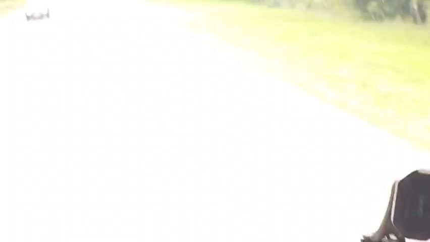 The video shows a view from a vehicle driving on a two-lane road. Trees line both sides of the road. There are other vehicles on the road, including a car in the left lane and a truck in the right lane ahead. The weather appears to be overcast. There are some road signs visible on the right side of the road.