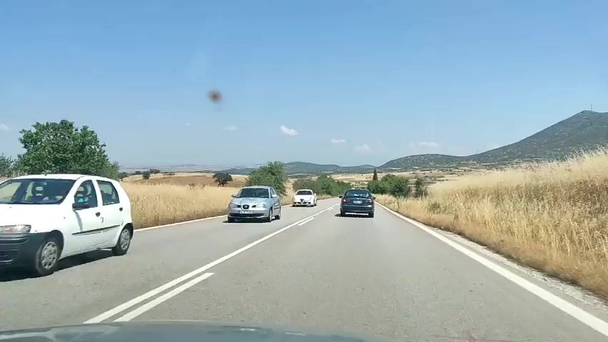 A first-person view from a vehicle shows a blue car and a white car driving on a two-lane road in a rural area. The camera is positioned behind the blue car, which is in the left lane. The white car passes the blue car and moves into the right lane. The camera then shows the blue car in the left lane, and the white car has moved ahead. The road is surrounded by dry, yellow fields and hills in the distance. The sky is clear and blue.