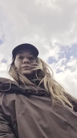 A woman with long brown hair and a black baseball cap, wearing a brown jacket, is the focus of the video. She stands on a rocky, elevated spot. In the background, there's a mountain range under a cloudy sky. A metal railing is visible behind her, overlooking a valley with some buildings. She initially looks at the camera, then gazes off to the side, and then back at the camera, giving a slight smile.