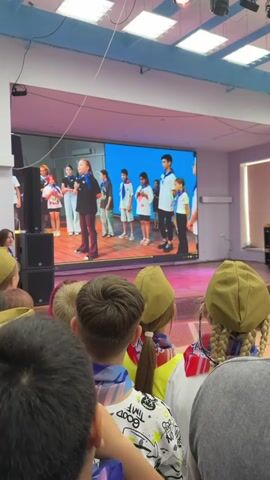 The video shows a group of children on a stage in front of a large screen displaying a performance. The children on stage are wearing white shirts and blue scarves. One child in the center is speaking into a microphone. In the foreground, the backs of the heads of several audience members are visible, some wearing green hats and blue scarves. The environment appears to be an indoor venue with stage lighting and speakers. The screen behind the performers displays text in Russian and images of children in performance.