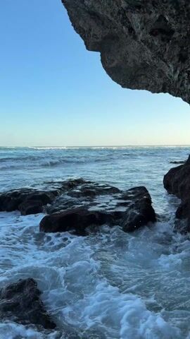 The video shows a view of the ocean from under a rock overhang. The sky is blue, and the ocean is turquoise with small waves. Dark rocks are visible in the foreground, with the waves crashing around them. The camera is static.