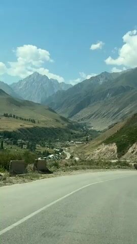 The video captures a scenic mountain landscape with a winding road in the foreground. The road curves to the right, with a paved surface and white lane markings. On the left side of the road, there is a concrete barrier and a grassy embankment. Further back, a valley unfolds, dotted with trees and small buildings, suggesting a settlement. Majestic mountains rise in the background, their peaks reaching towards a clear blue sky with scattered white clouds. The slopes of the mountains are covered in a mix of green vegetation and rocky terrain. The overall impression is one of natural beauty and a sense of journey through a mountainous region.