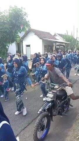 The video shows a procession of people in matching navy blue and white outfits marching down a street. They appear to be participating in some kind of parade or cultural event. A man on a motorcycle with a number 202 on its side rides in front of the group, leading the way. The houses lining the street are simple and appear to be in a rural setting. Some of the participants are holding what look like small flags or banners. The overall mood seems festive and communal.