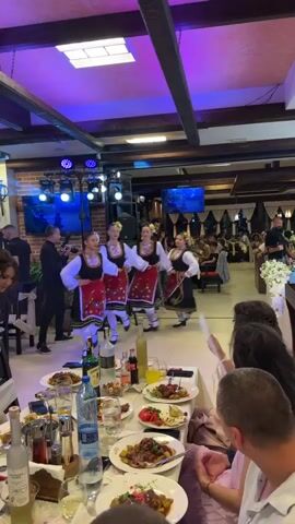 A vertical video shows people dancing and eating at a banquet hall. Four women in traditional Bulgarian folk costumes dance in a circle, holding hands. They wear black skirts with red and white embroidered aprons, white long-sleeved blouses, and black vests with colorful embroidery. Their hair is braided and adorned with red ribbons and flowers. One woman in a white, off-the-shoulder dress approaches them from the left and starts dancing with them. A band with a male singer performs in the background. Several tables are set with white tablecloths, plates of food, drinks, and wine bottles. Guests are seated around the tables, watching the dancers and enjoying their meals. The room has wooden beams on the ceiling, brick walls, and purple and blue lighting. Two large screens display images of water or abstract patterns.