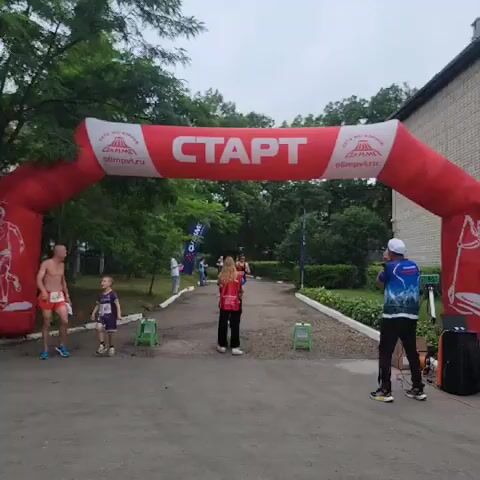 A man in a black and red running outfit with sunglasses and a race bib crosses a finish line under an inflatable arch that says START in Russian. A woman in a red vest and black pants stands to his left, appearing to assist him. Another runner in an orange outfit and a child in a purple shirt are visible to the left of the arch, as is a person in a blue and white shirt standing on the right side of the image, facing away from the camera. Several flags and some equipment are also visible. The scene appears to be outdoors in a park-like setting with trees and greenery.