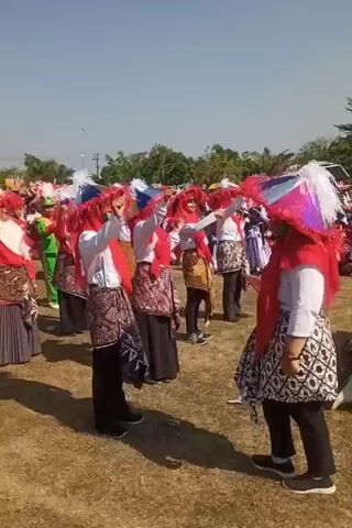 This is a medium shot video of a group of people in traditional dress, participating in an outdoor event. The people are wearing ornate headdresses in red, white, and blue, and red scarves. They are also wearing patterned skirts or sarongs over black pants. Some individuals are holding umbrellas or parasols. The setting appears to be a grassy field under a clear blue sky, with trees and buildings visible in the background. The participants seem to be engaged in a performance or parade, with some individuals moving and gesturing. The overall atmosphere appears festive and celebratory.