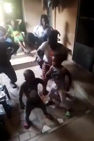 The video shows a lively gathering with music and dancing, featuring adults and children. The scene is set in what appears to be an indoor patio or covered area, with people sitting on chairs in the background. Several individuals are standing and dancing to music, with two young children particularly prominent in the foreground. One child is wearing a gray patterned outfit, and the other is in white. An adult is dancing with the children, guiding their movements. The camera angle shifts slightly, capturing more of the surrounding environment, including furniture, objects, and other people present at the gathering. The atmosphere appears joyful and celebratory.