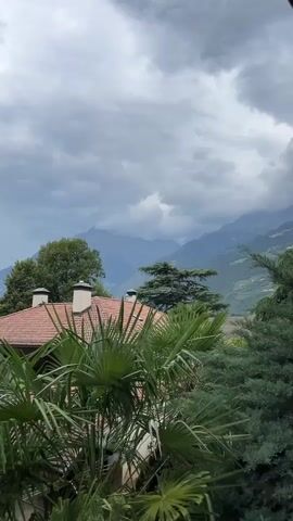 A slow-motion shot looks over foliage and a red-tiled roof towards a mountain range under a cloudy sky.