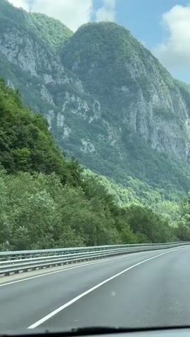 The video shows a view from inside a moving vehicle, looking out through the windshield at a scenic mountain landscape. A paved road stretches ahead, bordered by a guardrail on the left. Lush green trees cover the lower slopes of the mountains, which rise steeply in the background, displaying rocky cliffs and varied terrain. The sky is blue with scattered white clouds. The interior of the vehicle, including the dashboard and windshield, is visible in the foreground. The camera is relatively stable, indicating a smooth ride.