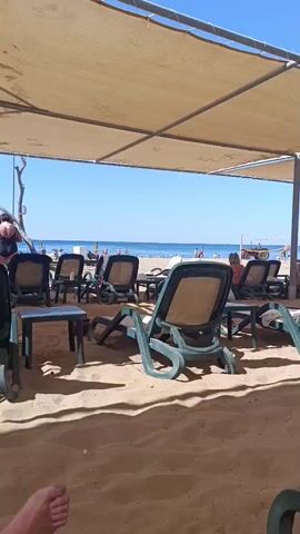 The video pans across a beach with rows of empty lounge chairs set up under a tan canopy. The chairs are mostly green plastic with tan fabric. The beach is covered in sand, and in the distance, the ocean is visible with a clear blue sky above. There are a few people visible in the distance on the beach and in the water. A volleyball net is set up on the beach. In the background, a lifeguard tower with a yellow flag is visible. There is also a red vehicle in the distance. The video seems to be taken from a seated position.