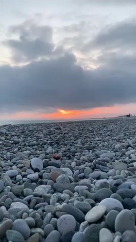 The image captures a pebble beach at sunset. The foreground is filled with smooth, gray, and white pebbles of varying sizes. In the distance, a thin strip of orange light pierces through the thick, gray clouds that dominate the sky. Two indistinct figures stand on the beach near the horizon. The overall atmosphere is calm and serene.