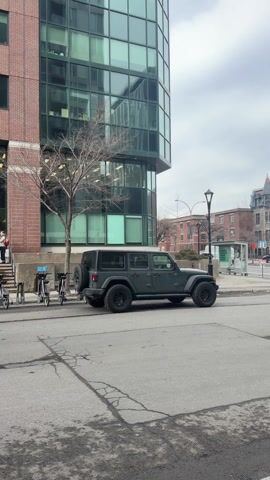 A red fire engine passes a grey Jeep Wrangler on a city street. A modern glass building is in the background behind the Jeep and a brick building is behind the fire engine. The sky is overcast.
