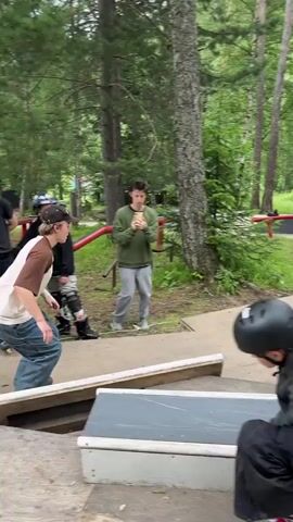 At 00:00, a person in a white and brown shirt and blue jeans is on roller skates, crouched down on a ramp. Other people are visible in the background, some standing and watching, others on skates. The environment appears to be an outdoor skate park with ramps, rails, and trees. At 00:01, the person on skates jumps and grinds on a red rail. At 00:02, the person continues to grind on the rail, arms outstretched for balance. At 00:03, the person finishes the grind and approaches another ramp. At 00:04, the person is on the ramp and appears to be preparing for another trick.