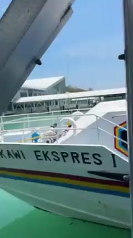 The video shows a boat dock with a white boat docked. The boat has blue trim, and a red, yellow, and blue design on the side. The camera follows three women walking along the dock towards the boat. They are all wearing backpacks. The dock has a metal structure with a white roof. The sky is blue and clear.