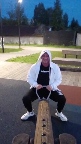 A person is sitting on a wooden seesaw, holding onto the handles. They are wearing a white hooded jacket, a black t-shirt with a logo, black sweatpants, and white sneakers. The person is smiling and appears to be in motion on the seesaw. The background shows an outdoor playground area with trees, a fence, and some seating. The lighting suggests it is dusk or evening.