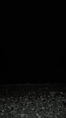 This is a short video of a beach at night. The foreground is filled with pebbles, and in the distance, the ocean is visible. The water is dark, and there are no lights or people present. The sky is completely black, indicating it is nighttime. The camera is positioned low to the ground, facing the ocean.