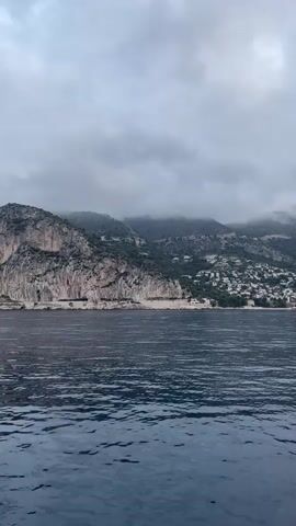 The video shows a view of the sea with a coastline and mountains in the background. The water is dark blue with small waves. On the coastline, there are many houses built on the hills. The mountains are steep and covered with trees. The sky is cloudy and grey. There are two large yachts visible in the distance on the water.