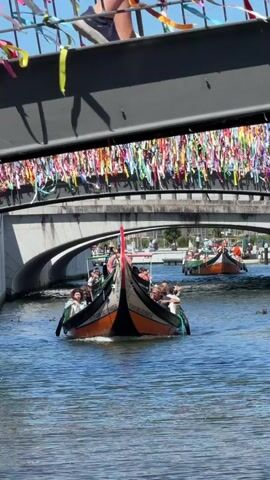 A gondola with many passengers travels down a river under a bridge decorated with ribbons. Another gondola is visible in the background. The water is blue and calm. The sky is clear and sunny.