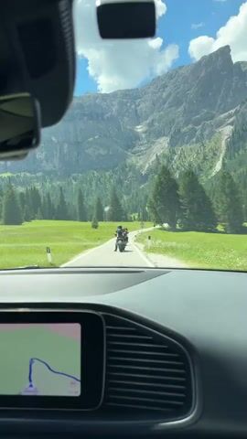 The video shows a view from inside a car dashboard with a navigation screen on the left side. The car is on a paved road, surrounded by green fields and pine trees. In the distance, a large, rocky mountain range is visible under a bright blue sky with white clouds. Several motorcycles are riding on the road ahead of the car, appearing to be part of a group or convoy. The scene suggests a scenic drive through a mountainous region.